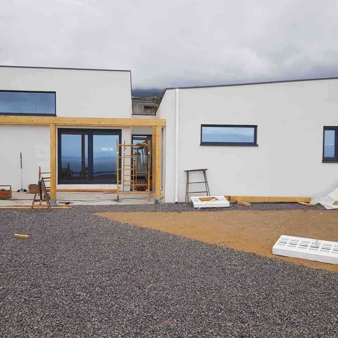 Exterior de una casa moderna en construcción con paredes blancas, ventanas azules y patio de grava.