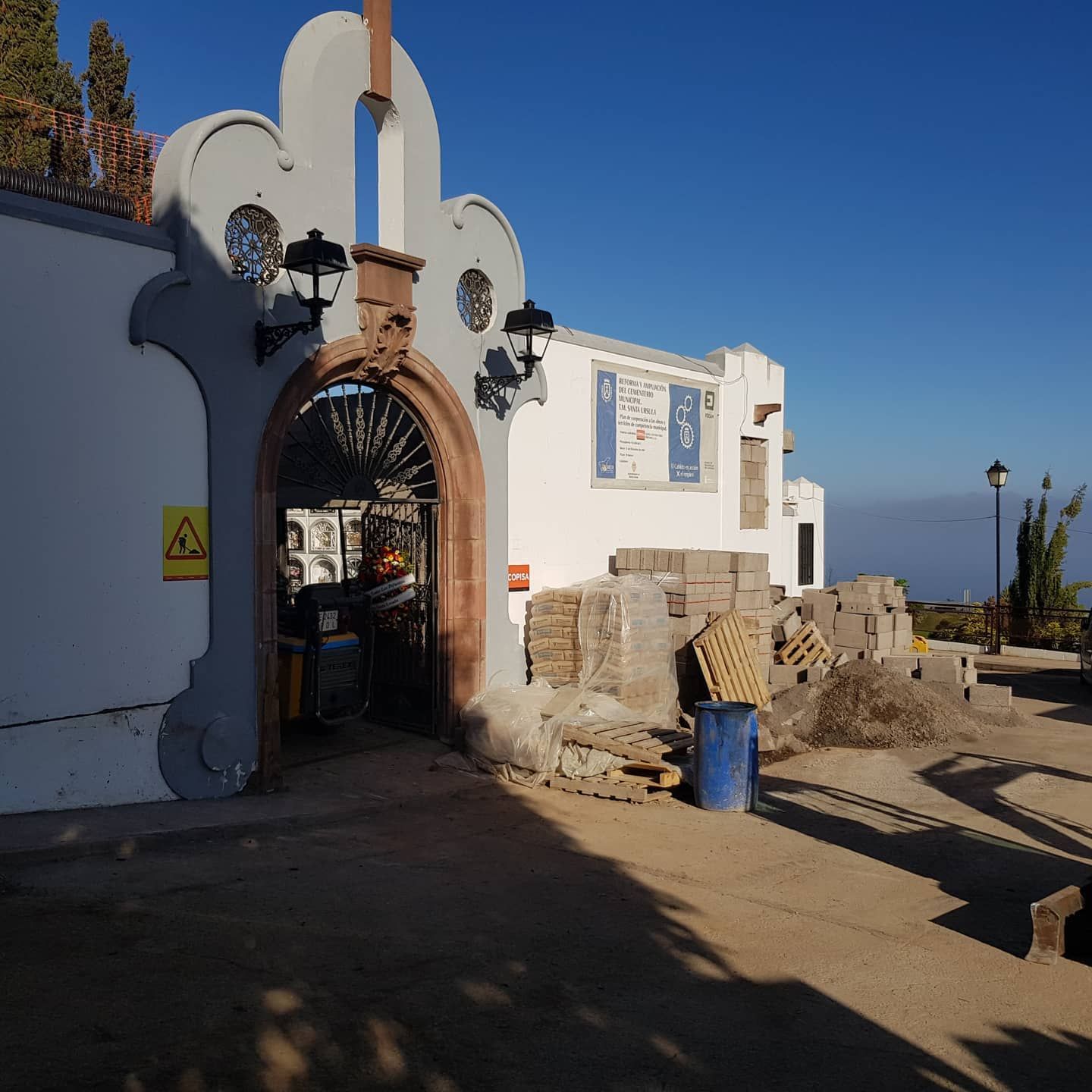 Entrada arqueada blanca a un edificio con faroles, escalones de piedra y un barril azul bajo un cielo despejado.