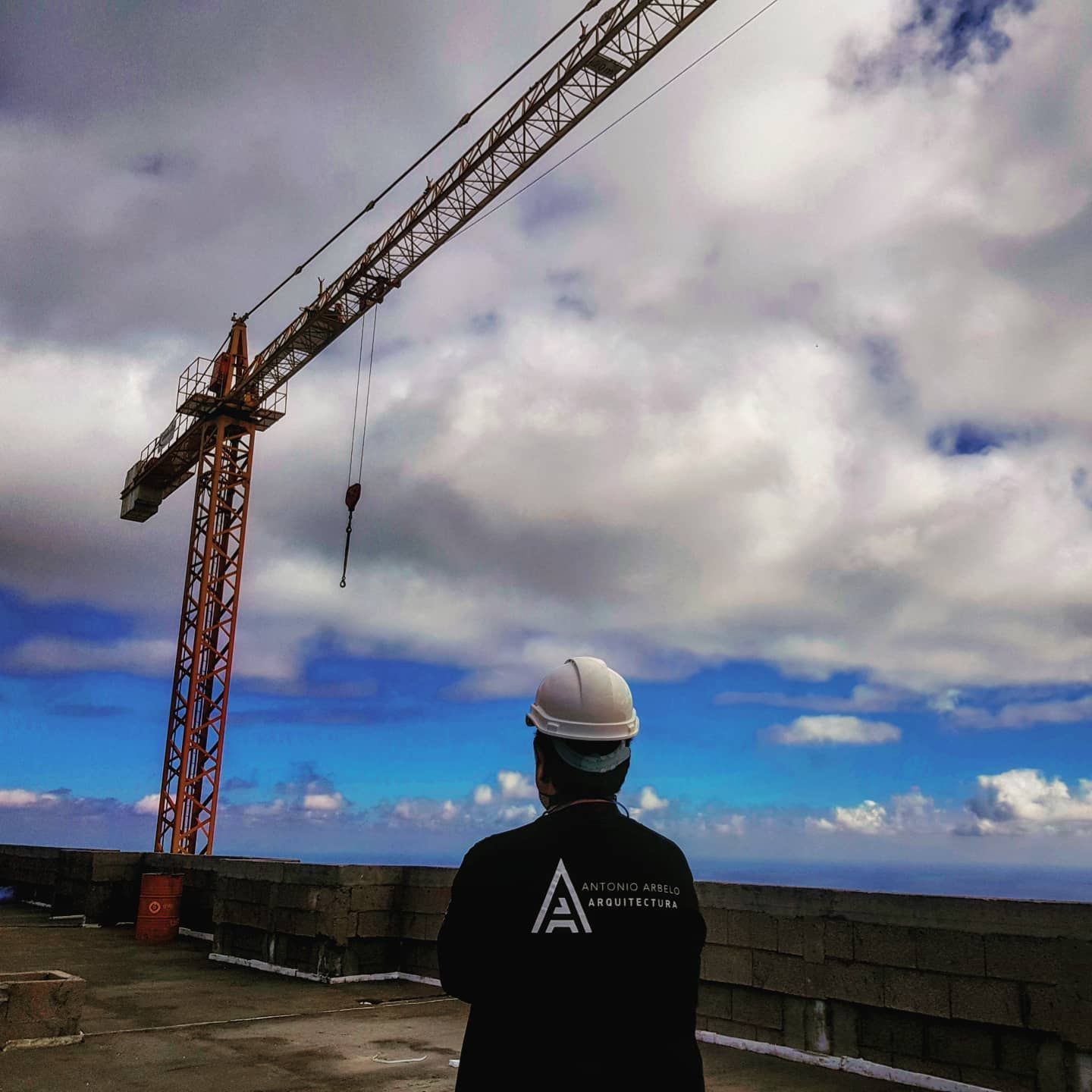 Un trabajador con casco observa una grúa en una azotea bajo un cielo nublado.
