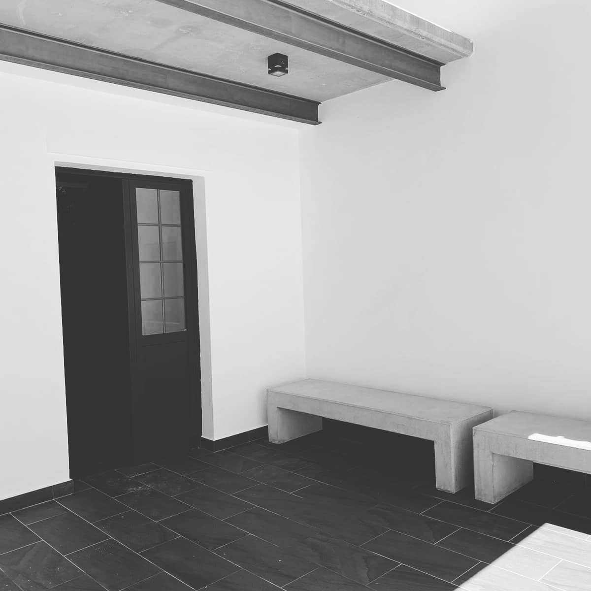 Habitación minimalista con suelo de baldosas oscuras, puerta negra, vigas de madera en el techo y bancos sencillos a lo largo de la pared.