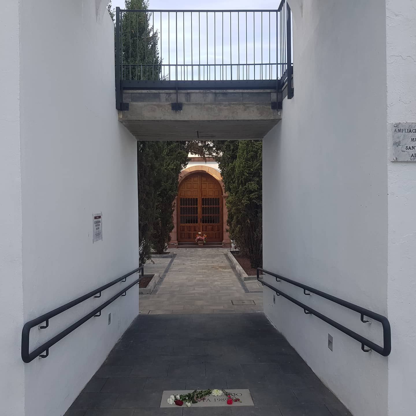 Un estrecho pasillo entre paredes blancas conduce a una puerta de madera con un pequeño balcón encima.