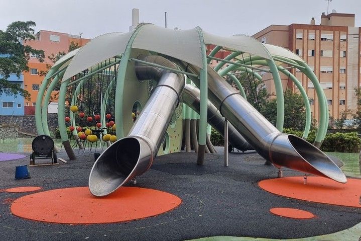 Parque infantil colorido con una estructura de escalada en forma de cúpula y dos toboganes metálicos brillantes sobre una superficie de goma.