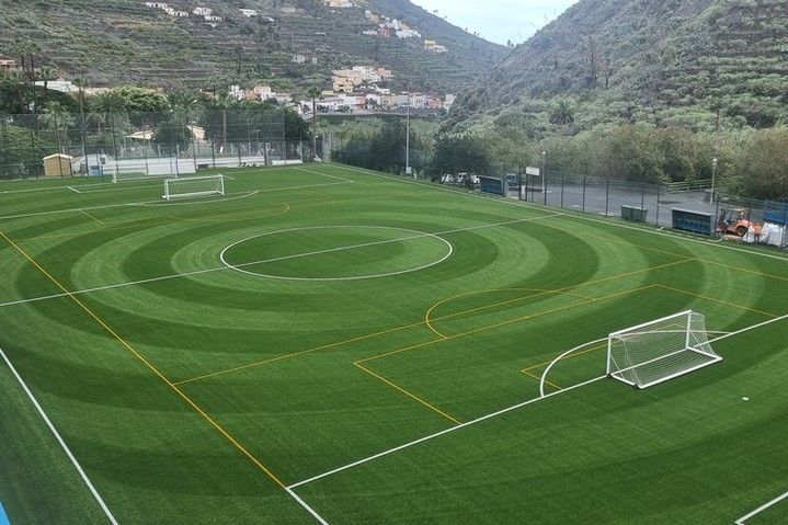 Campo deportivo verde vacío con porterías blancas y marcas circulares en un valle de montaña.