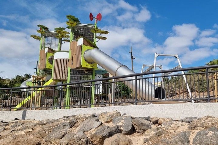 Parque infantil con un tobogán plateado alto, estructuras de escalada de madera y rocas en primer plano bajo un cielo azul.