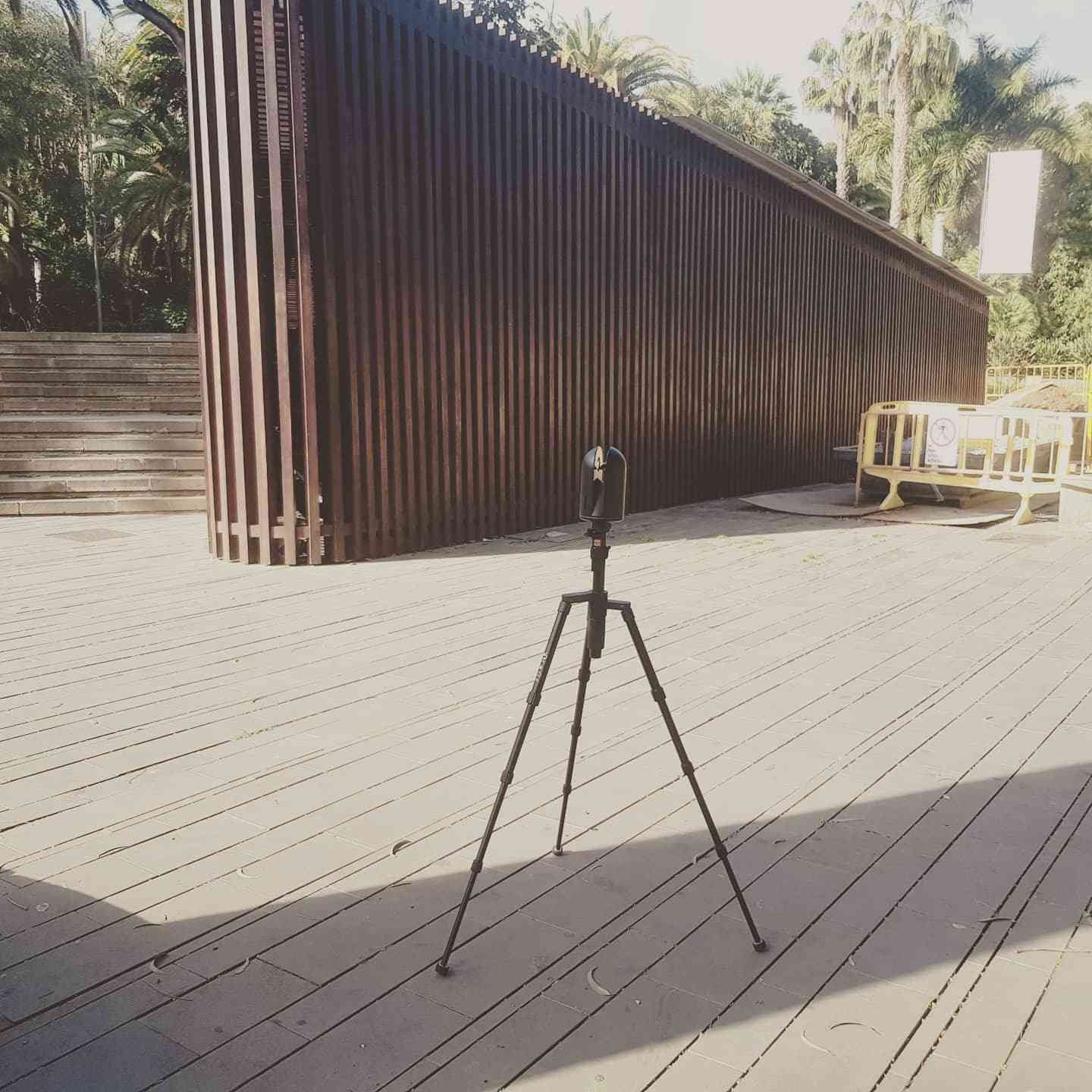 Trípode sobre una terraza de madera soleada junto a una estructura de metal corrugado y un banco.