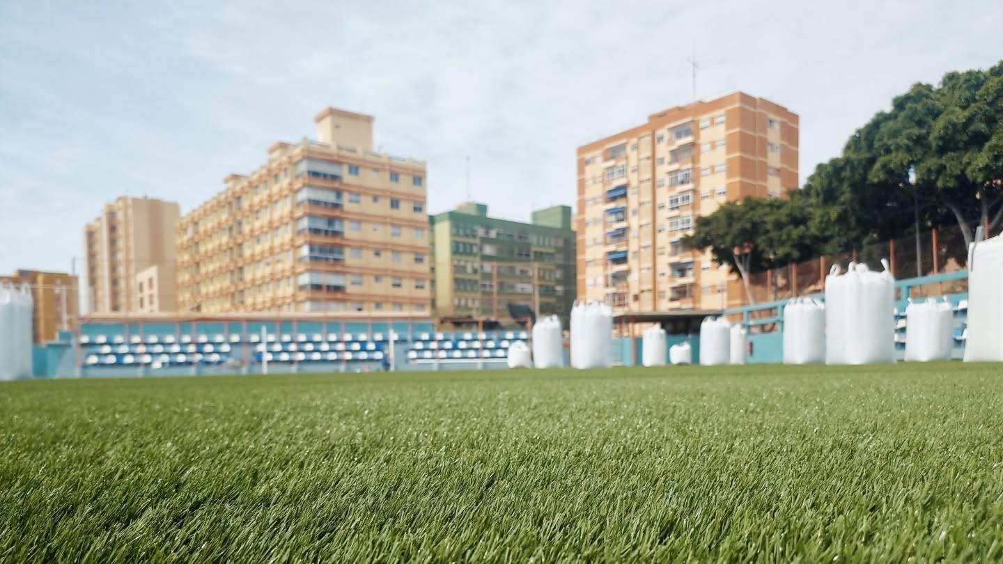 Campo deportivo cubierto de hierba con edificios de apartamentos naranjas y gradas blancas bajo un cielo brillante.