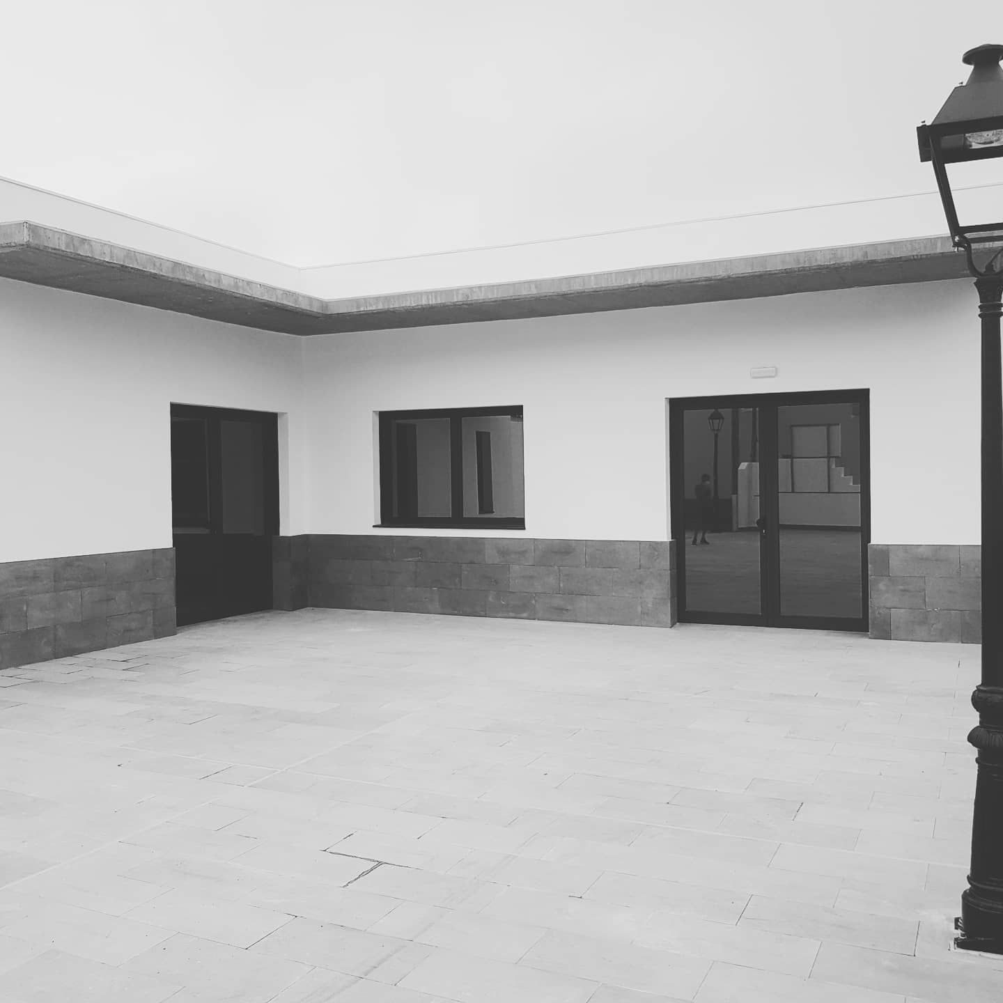 Patio vacío con baldosas, paredes blancas, puertas y ventanas oscuras y una farola a la derecha.
