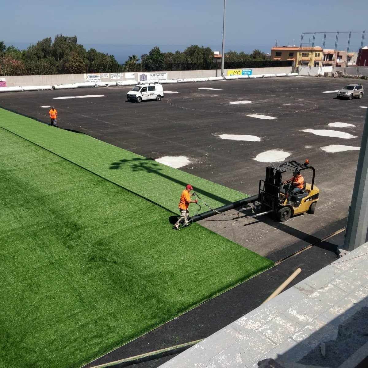 Trabajadores colocando césped artificial verde junto a un terreno pavimentado, con una carretilla elevadora y un camión blanco cerca.