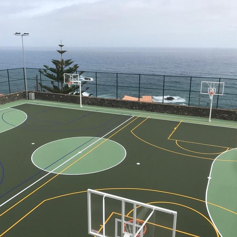 Cancha de baloncesto al aire libre vacía junto al océano, con líneas y aros pintados de verde y amarillo.