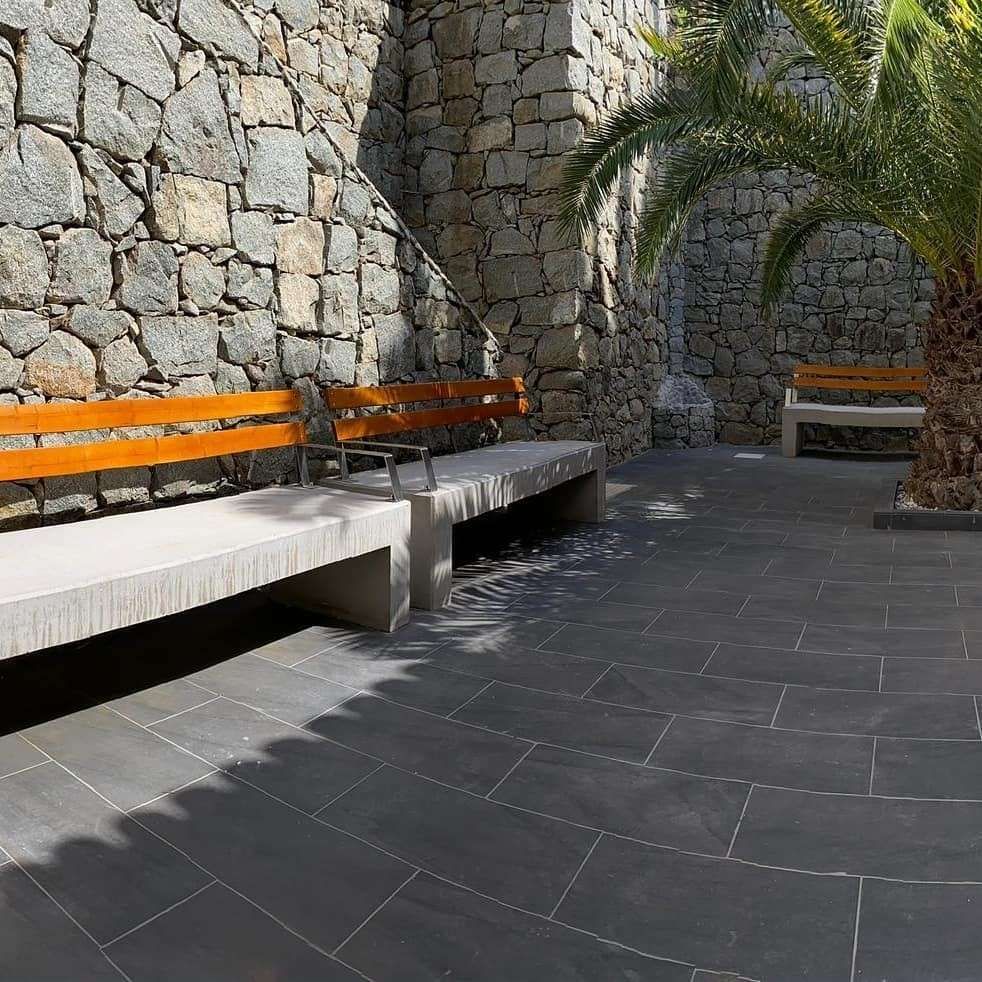 Patio de piedra con bancos naranjas, suelo de baldosas oscuras y una palmera bañada por el sol.
