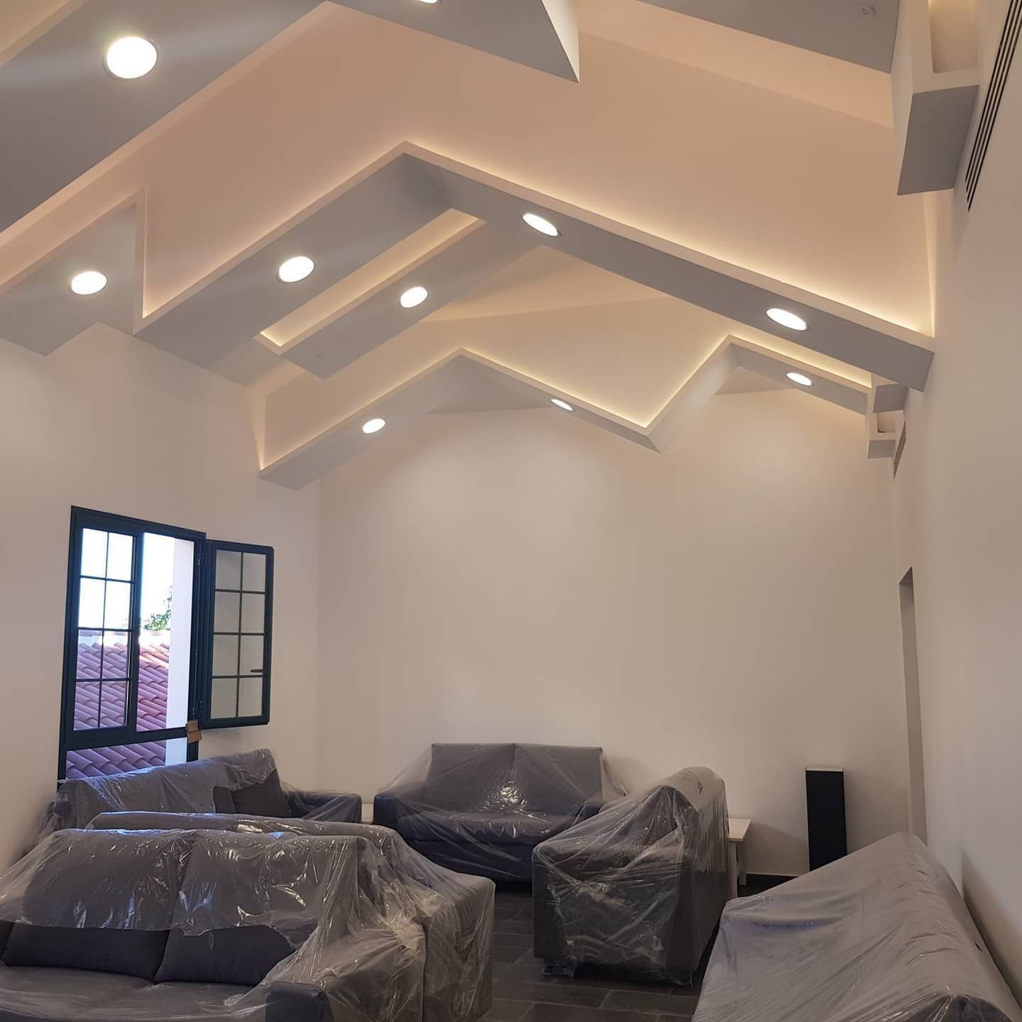 Sala de estar vacía con lámparas de techo inclinadas, muebles tapizados y una ventana abierta a la izquierda.