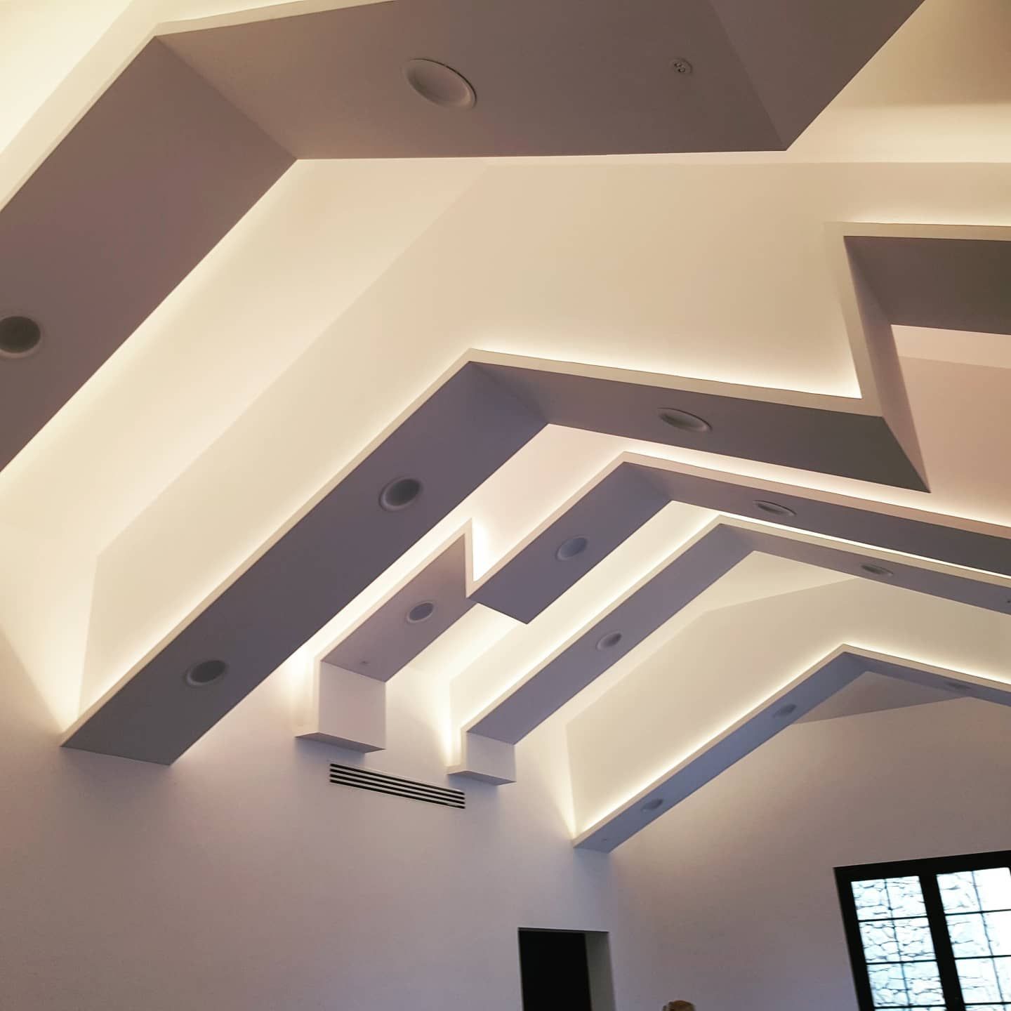 Techo geométrico empotrado con vigas blancas y grises en capas y luces redondas sobre la ventana de una habitación.