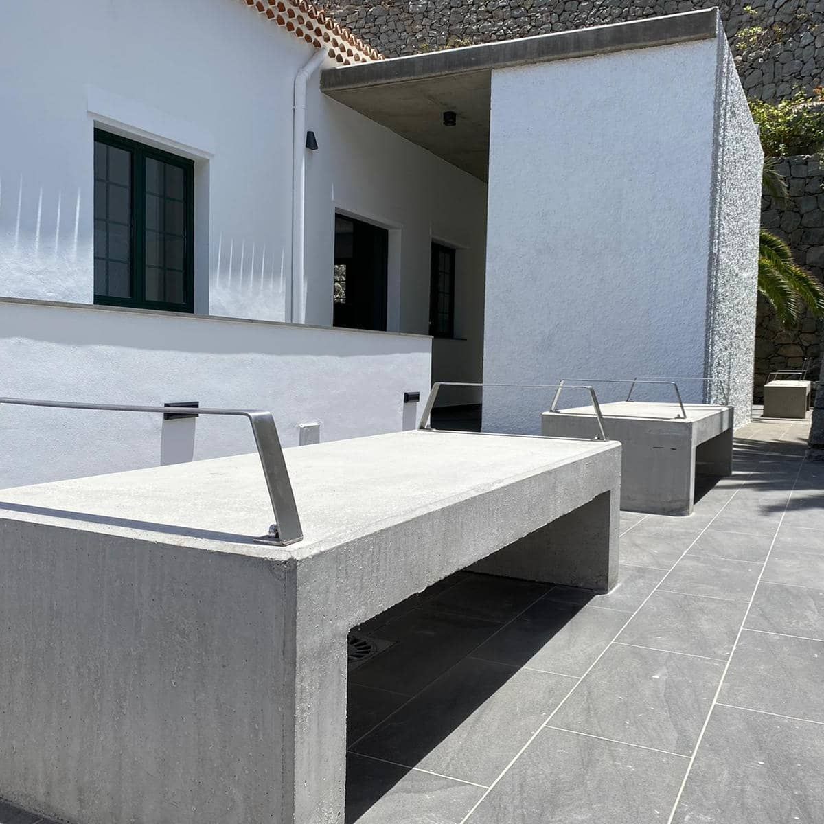 Patio exterior con mesas y bancos de hormigón junto a un edificio de estuco blanco bajo la luz del sol.