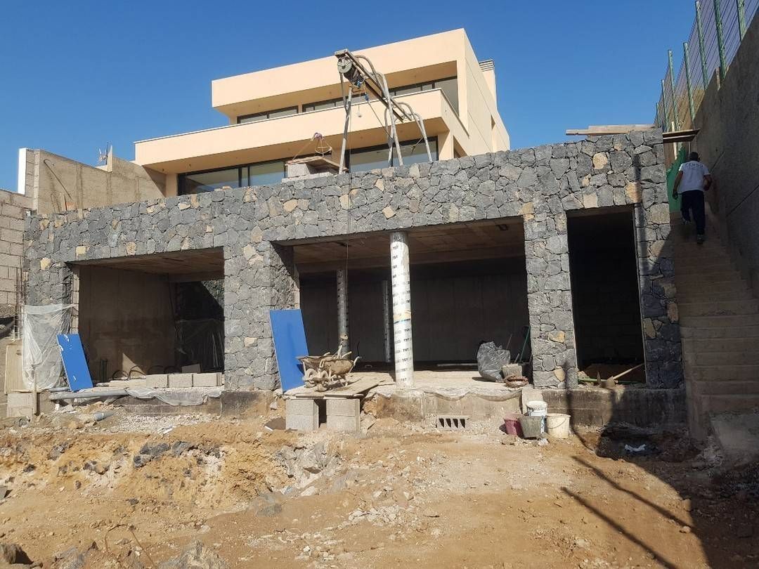Casa de piedra parcialmente construida con fachada abierta, terreno de escombros y un edificio beige detrás bajo un cielo despejado.