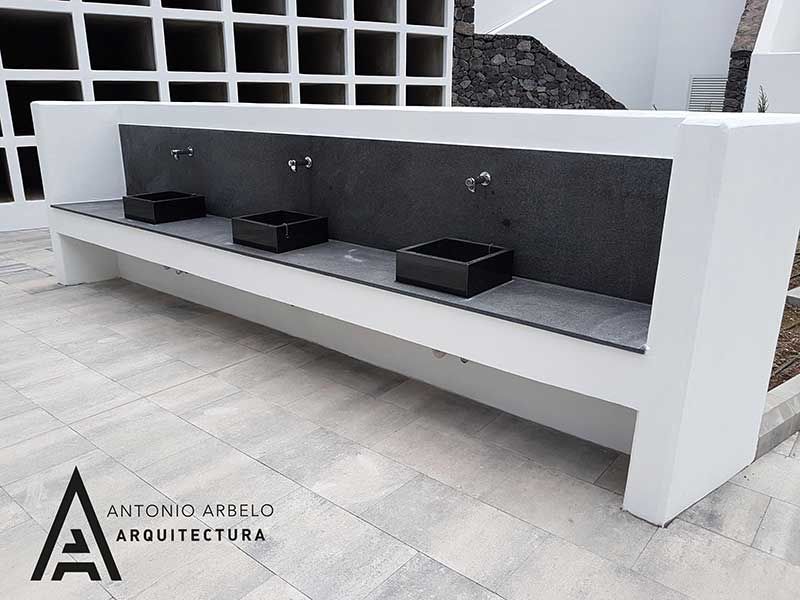 Banco moderno blanco para exteriores con respaldo negro y tres macetas negras en un patio embaldosado.