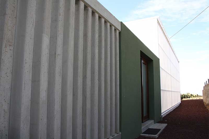Exterior de edificio moderno con pared gris acanalada, panel de entrada verde y pared lateral blanca a lo largo de un camino estrecho.