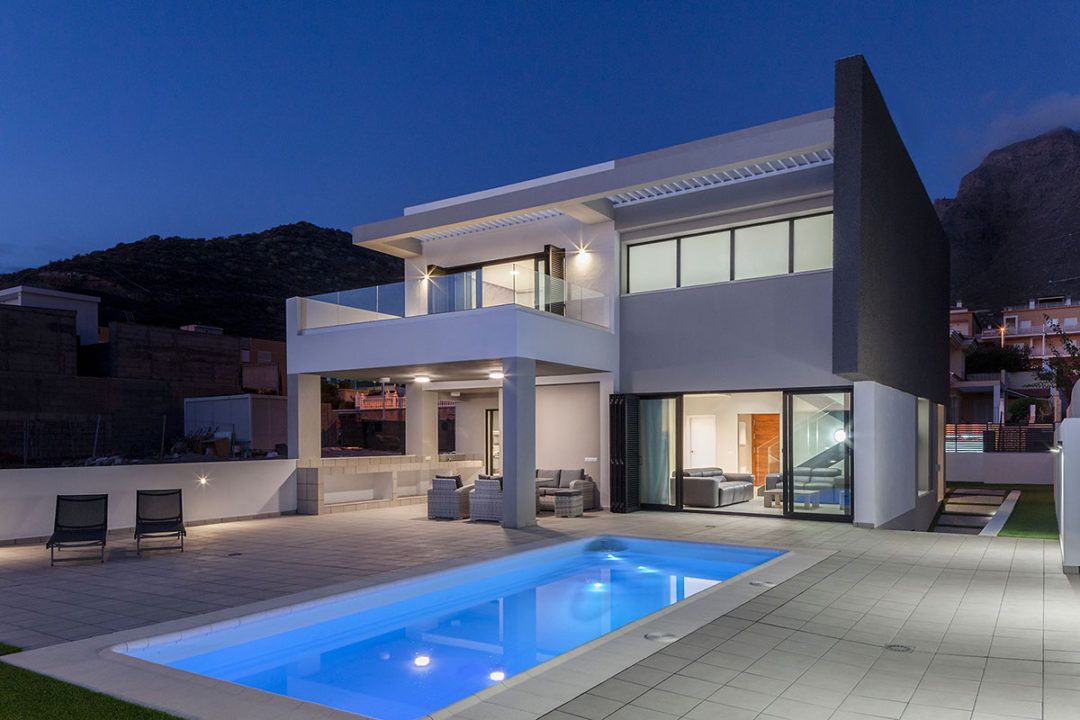 Moderna casa de dos plantas al atardecer con piscina iluminada y patio con vistas a una ladera.