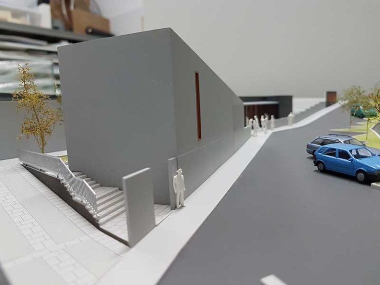 Maqueta de un edificio moderno de color gris junto a una calle con coches azules aparcados y pequeñas figuras.