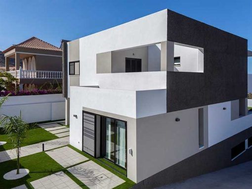 Moderna casa de dos plantas, blanca y negra, con balcón, puertas de cristal y jardín paisajístico bajo un cielo azul.