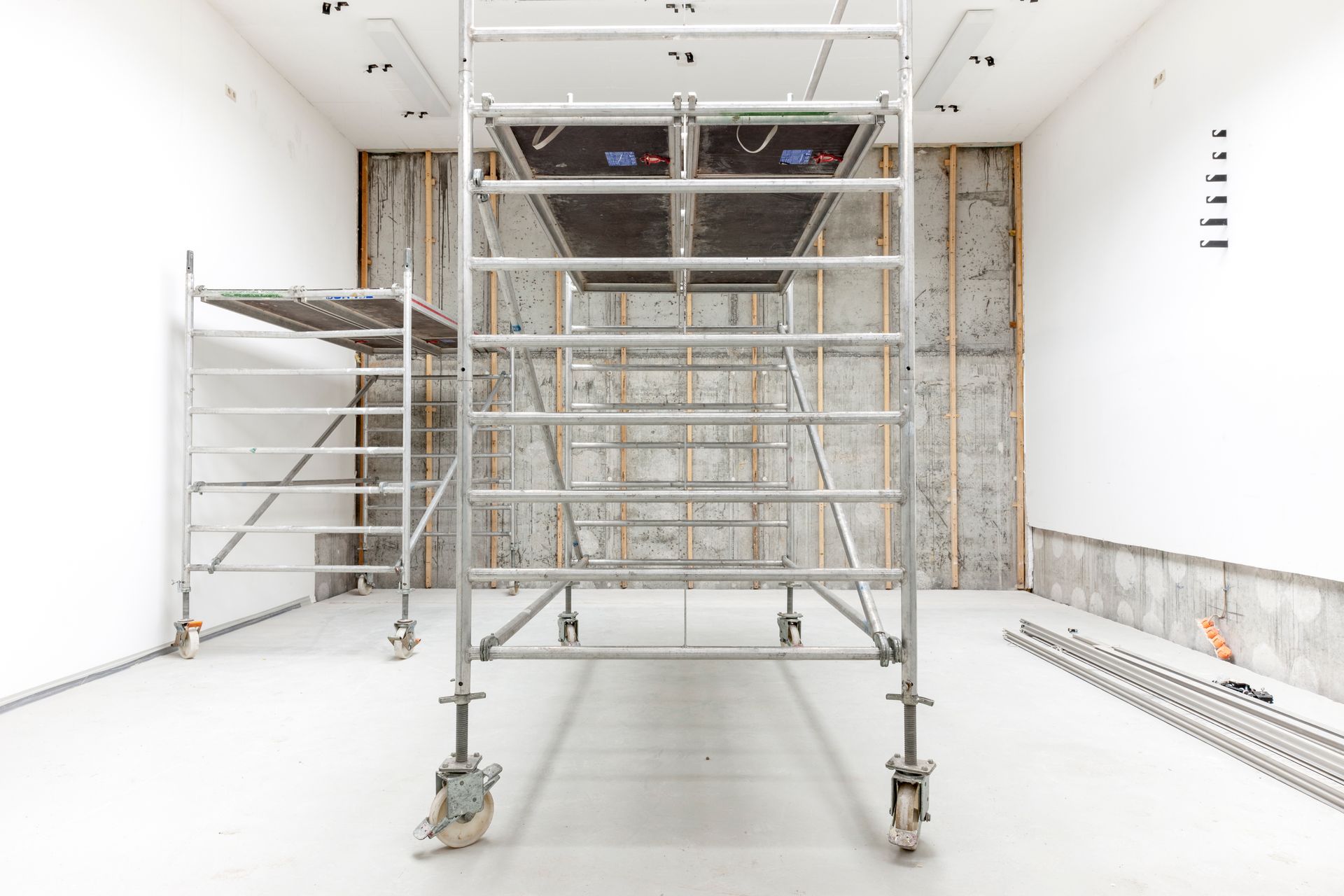 Échafaudage métallique à l'intérieur d'une pièce blanche, avec un mur partiellement apparent et des matériaux de construction.
