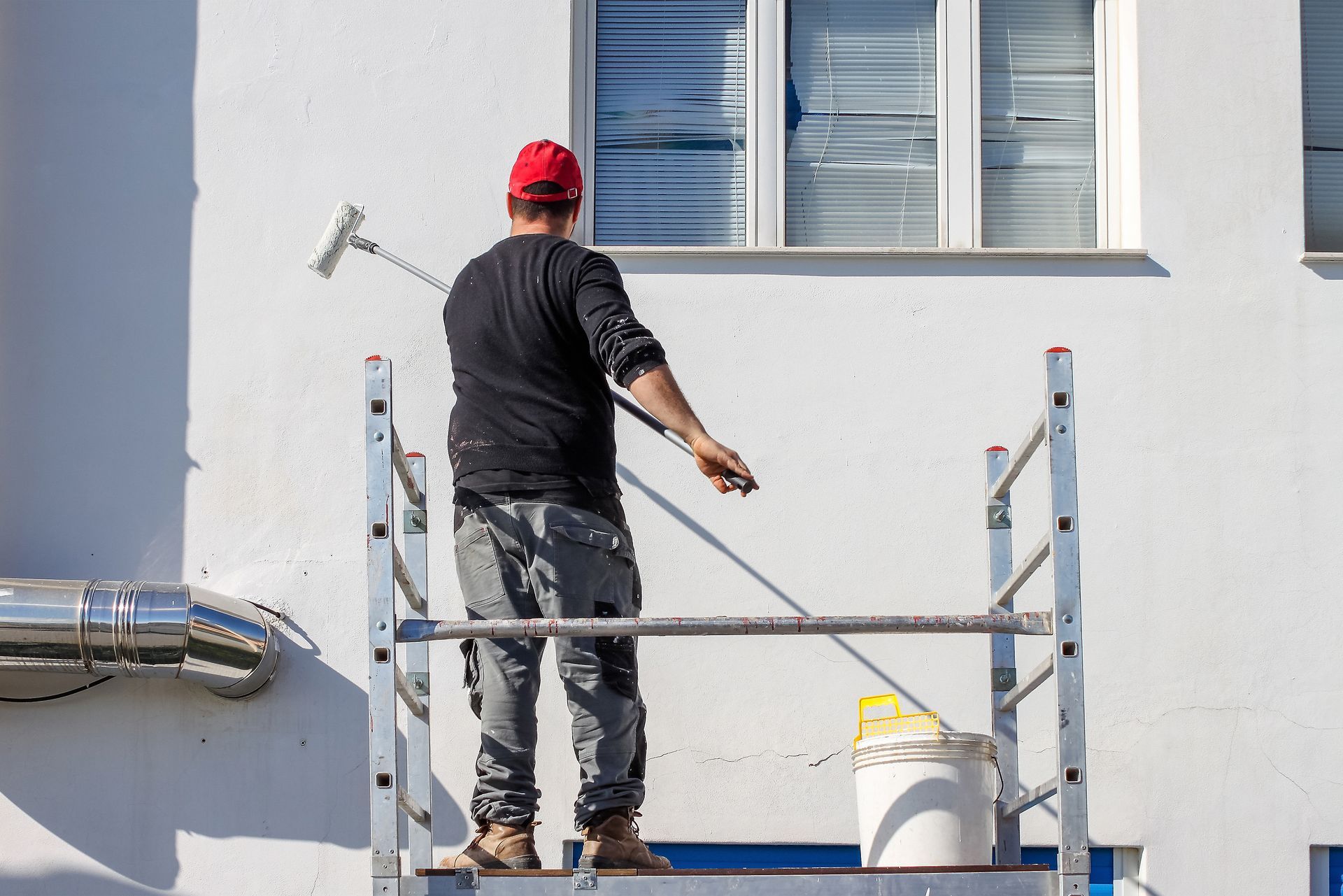 Un ouvrier peint la façade blanche d'un bâtiment depuis un échafaudage, à l'aide d'un rouleau et d'un pot de peinture.