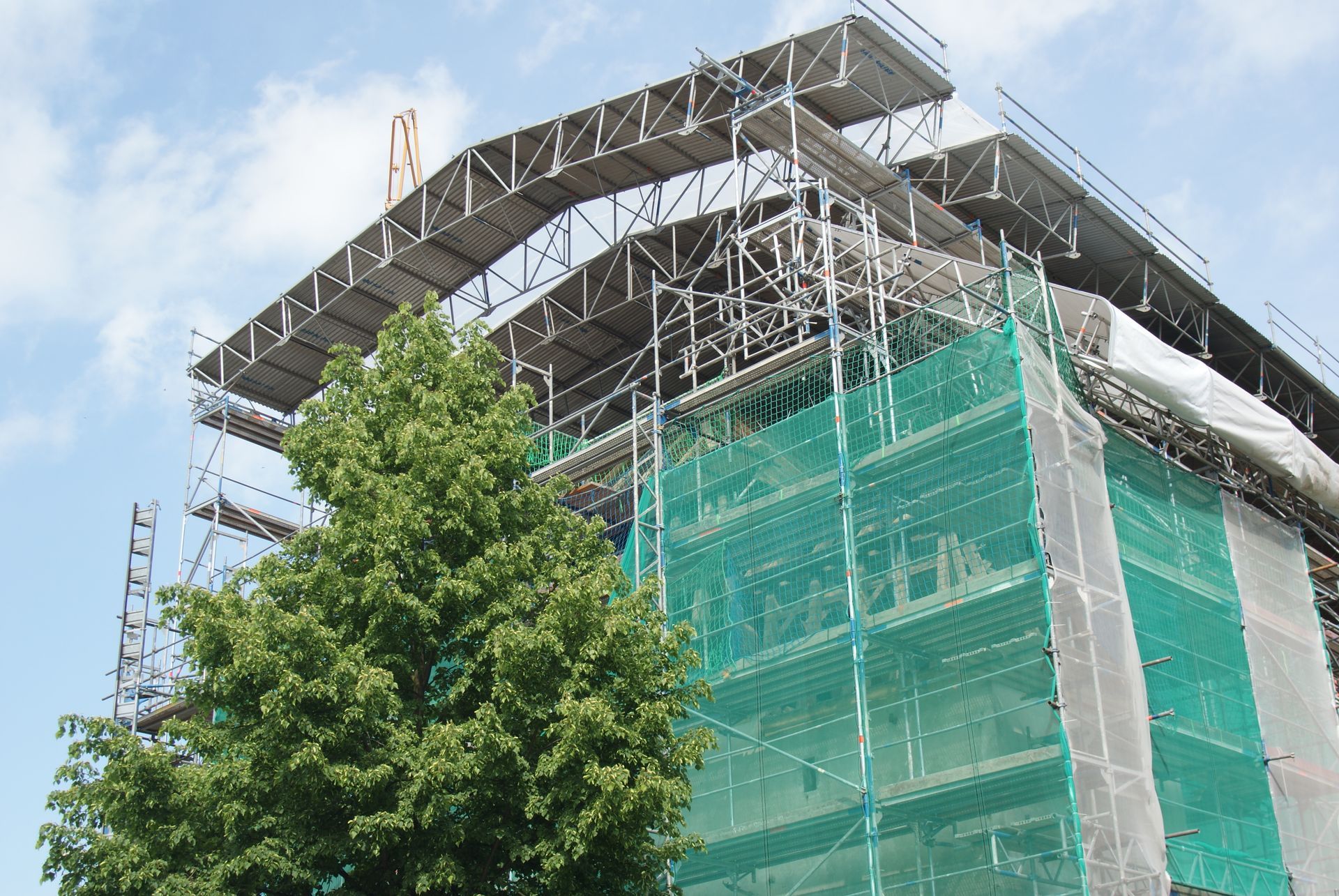Échafaudage sur un bâtiment avec filet de sécurité vert, un arbre au premier plan et un ciel bleu.