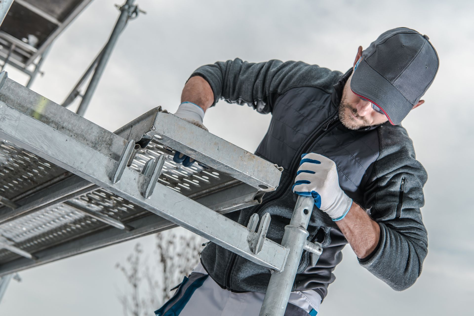 Un homme portant des gants et une casquette de travail assemble un échafaudage à l'extérieur.