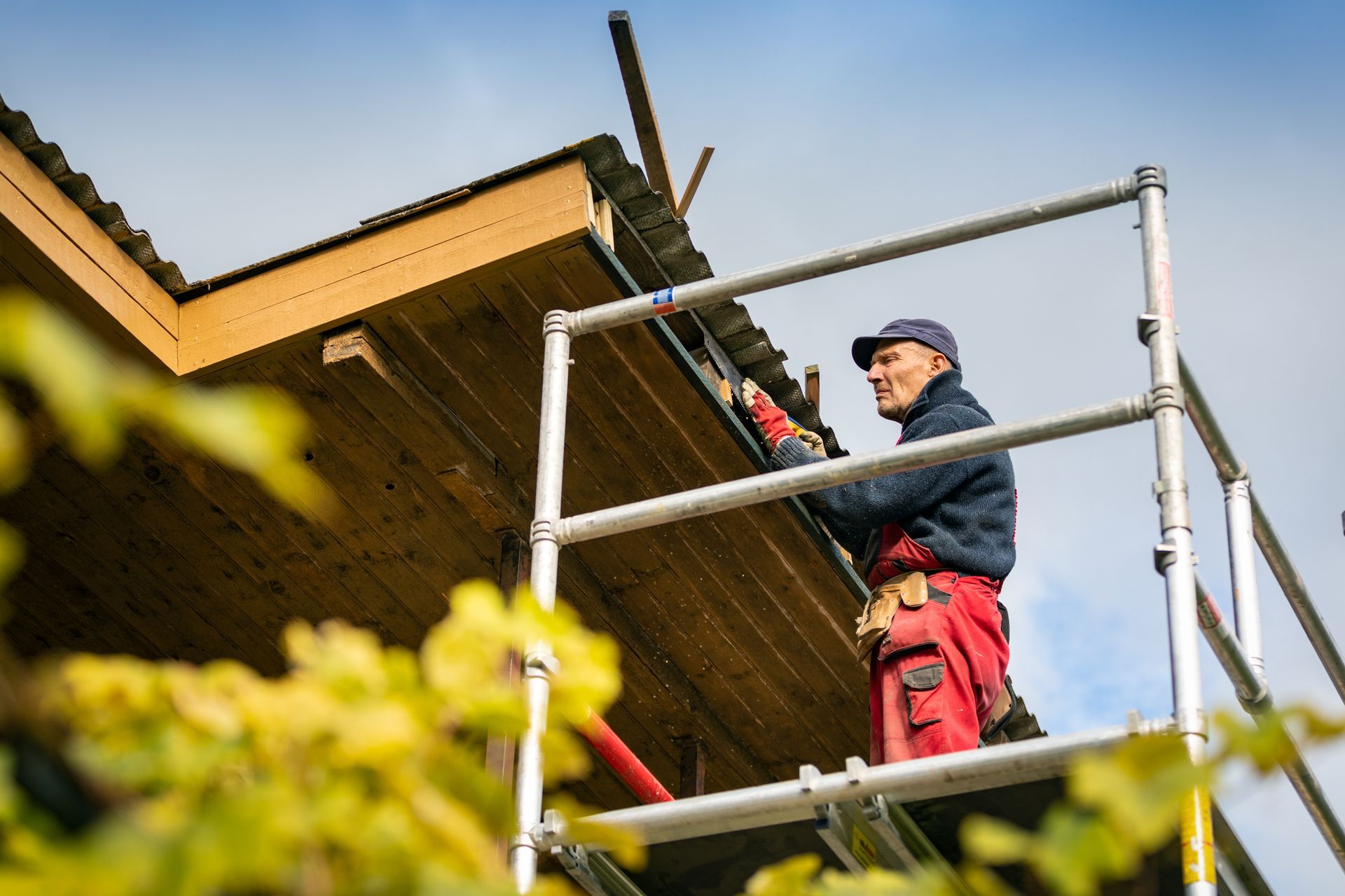 Ouvrier du bâtiment sur un échafaudage, installant des éléments de toiture. En extérieur, ciel bleu, pantalon rouge, ceinture à outils.