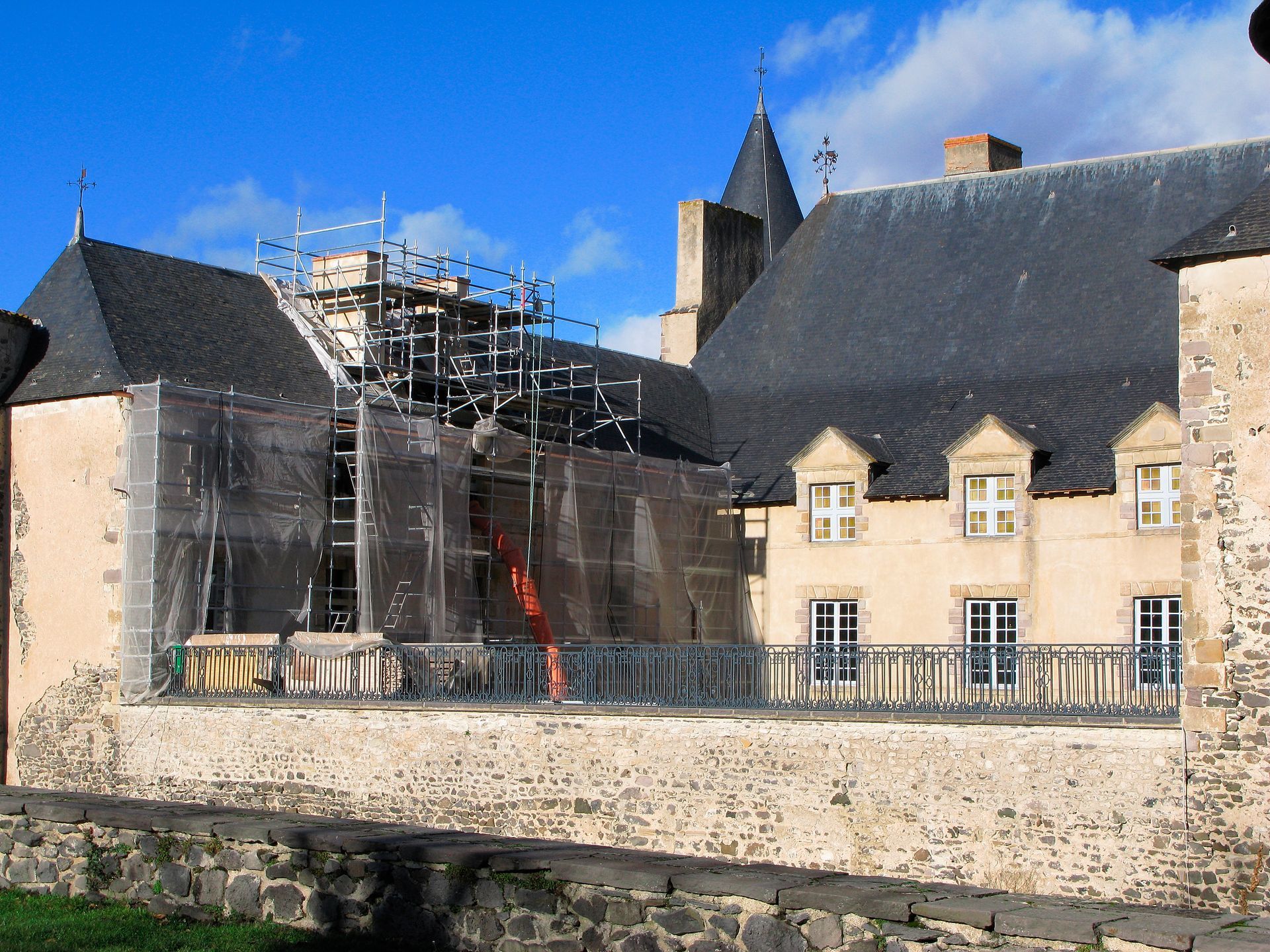 Bâtiment en pierre en rénovation, avec des échafaudages à côté d'un mur au bord de la rivière, sous un ciel bleu.