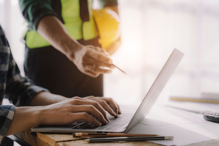 Une personne tape sur un ordinateur portable, un ouvrier du bâtiment montre du doigt, des plans et un casque de chantier sont visibles.