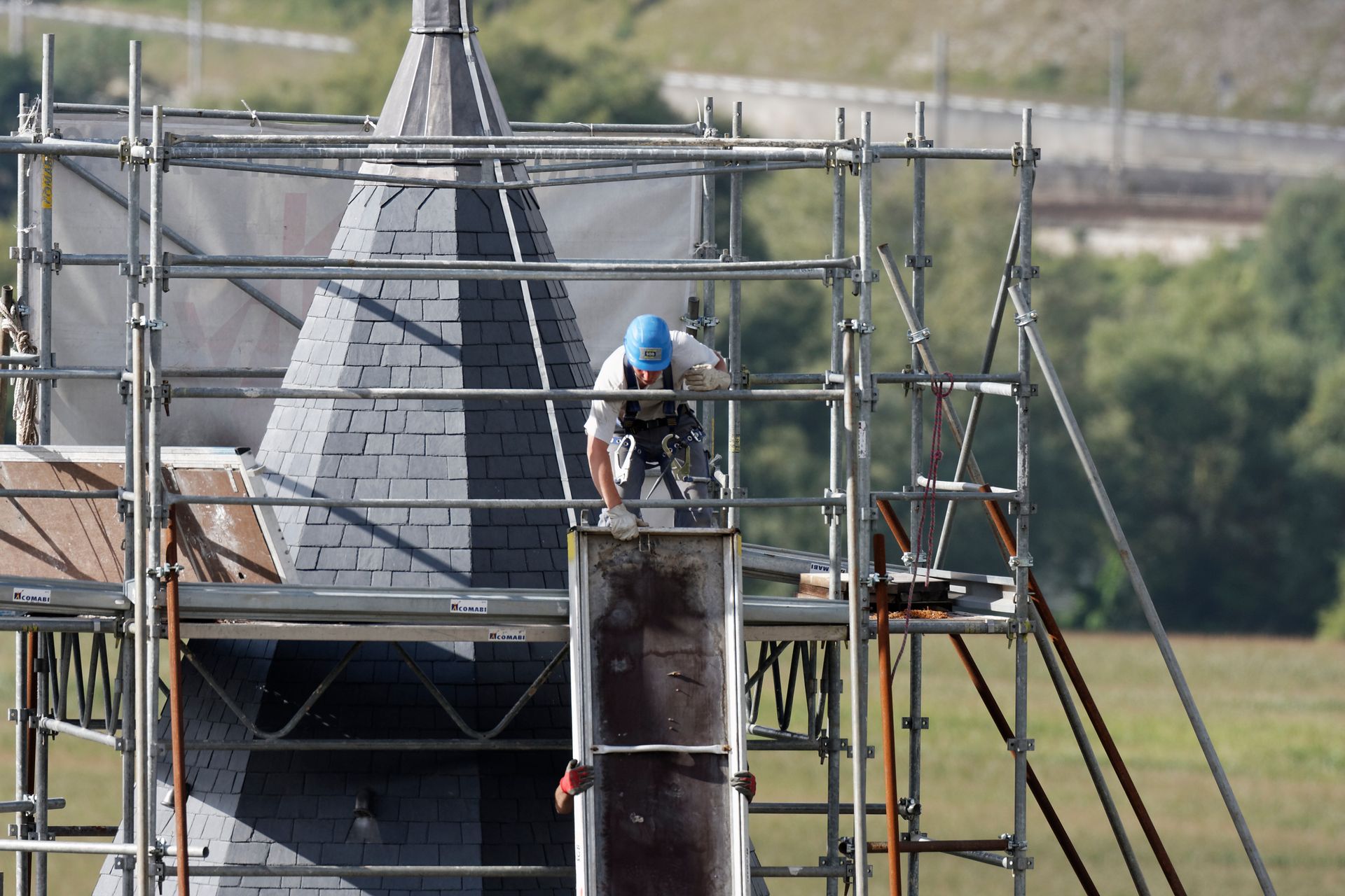 Un ouvrier répare un clocher sur un échafaudage. Tuiles grises, casque bleu, paysage rural.