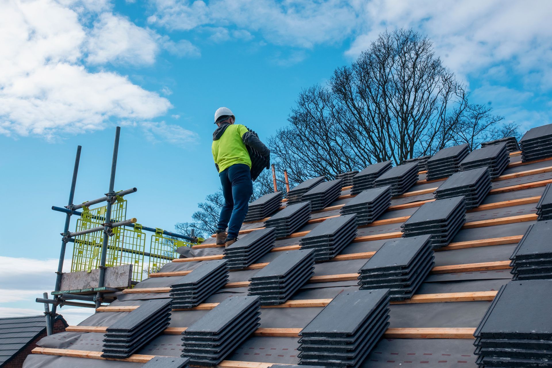 Un couvreur, portant un gilet de sécurité et un casque de chantier, transporte du matériel sur un toit de tuiles, avec des échafaudages et un ciel bleu.