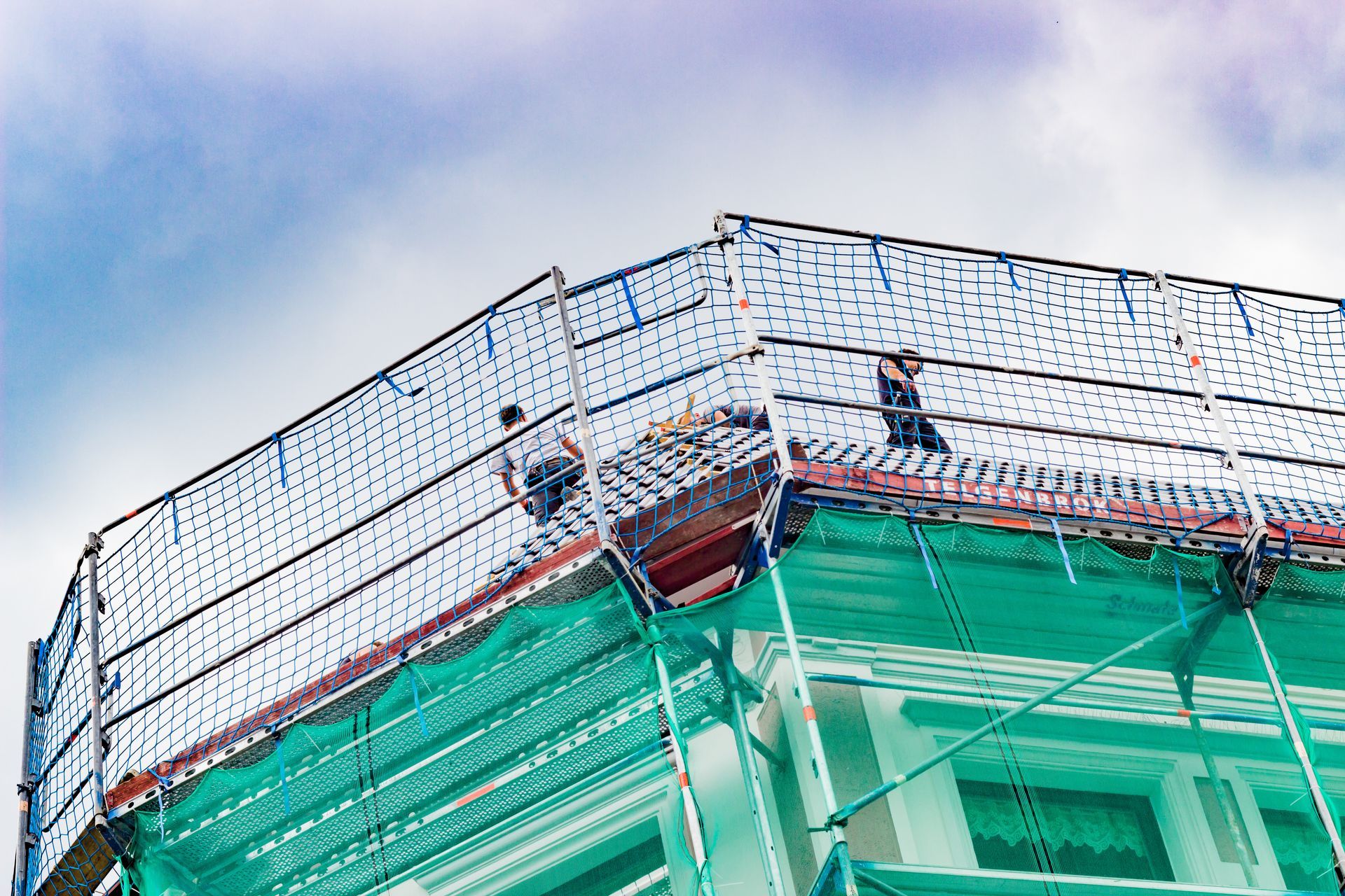 Des ouvriers du bâtiment travaillent sur le toit d'un immeuble, derrière des filets de sécurité. On aperçoit le ciel bleu et les filets verts.