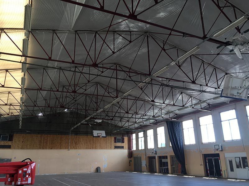 Vue intérieure d'un gymnase avec un haut plafond métallique, des paniers de basket et des fenêtres.