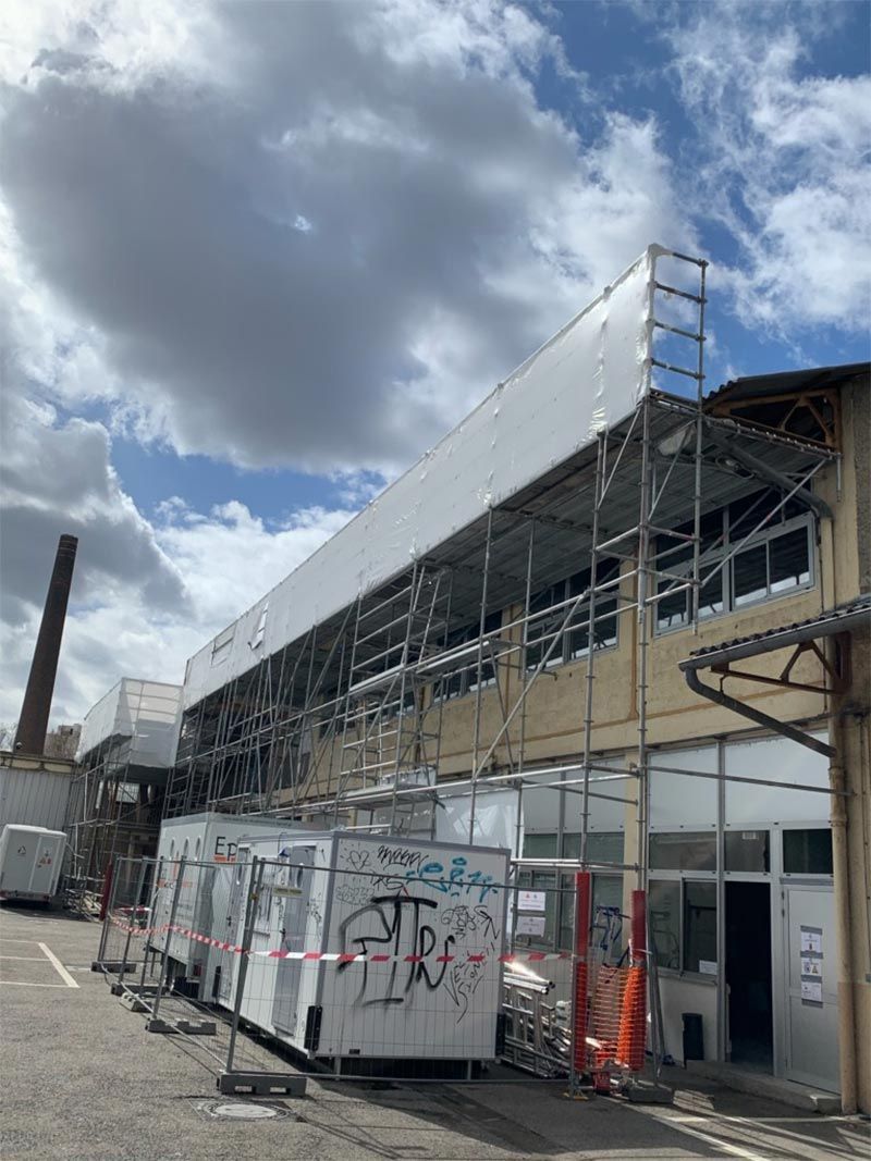 Des échafaudages recouvrent la façade d'un bâtiment ; des unités mobiles sont installées en dessous. Journée ensoleillée, ciel nuageux.