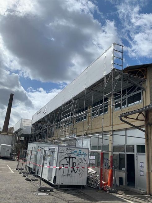 Des échafaudages recouvrent la façade d'un bâtiment ; des unités mobiles sont installées en dessous. Journée ensoleillée, ciel nuageux.