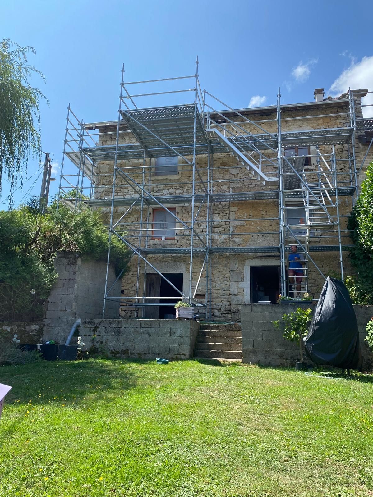 Échafaudages sur la façade d'une maison en pierre de deux étages, probablement en cours de rénovation. Journée ensoleillée, jardin verdoyant.