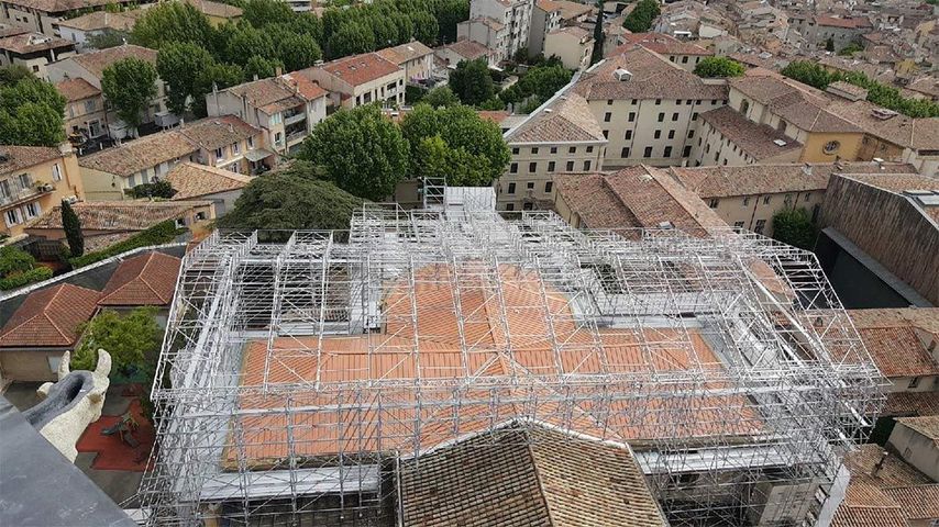 Vue en hauteur d'un vieux bâtiment en construction. Des échafaudages recouvrent le toit, entouré de toitures en tuiles de terre cuite.