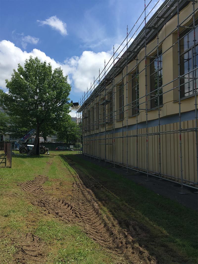 Vue extérieure d'un bâtiment en rénovation avec échafaudages, bardage jaune pâle et pelouse verte.