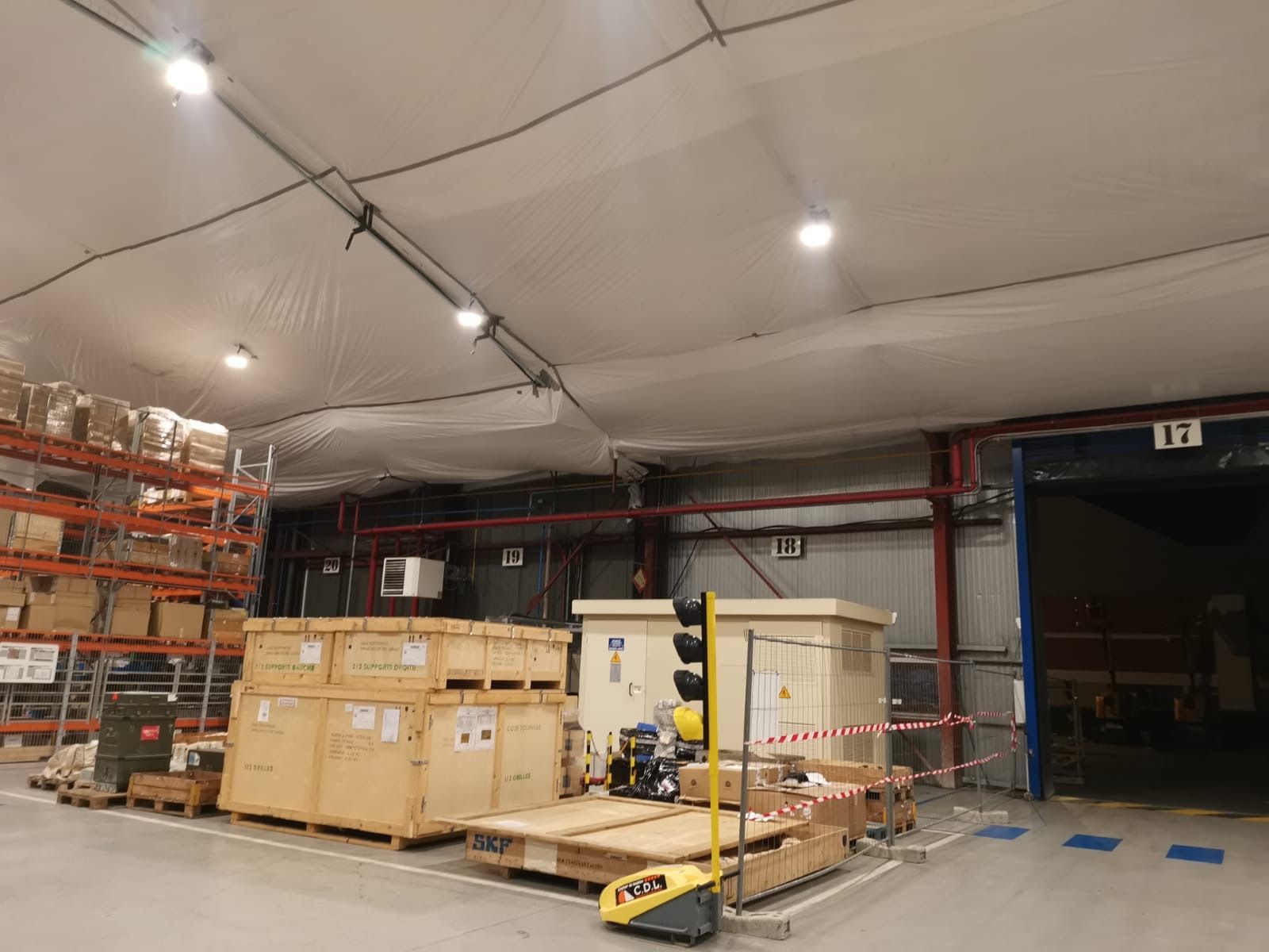 Intérieur d'un entrepôt avec caisses en bois, étagères et quai de chargement. L'éclairage et le matériel de sécurité incendie sont visibles.