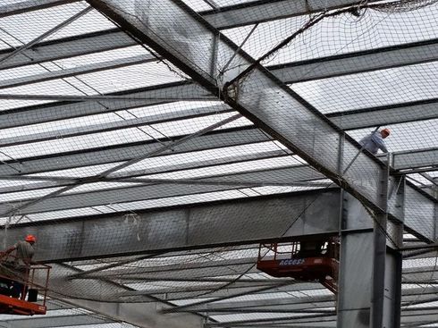 Des ouvriers sur des nacelles élévatrices installent des filets de sécurité sous les poutres d'acier du toit d'un bâtiment.