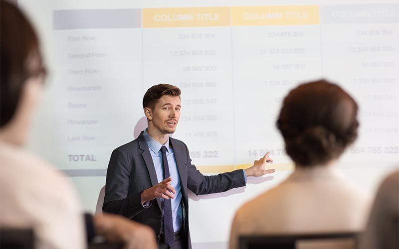 Un homme en costume s'adresse à un public en désignant une table projetée.