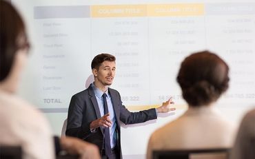 Un homme en costume s'adresse à un public en désignant une table projetée.