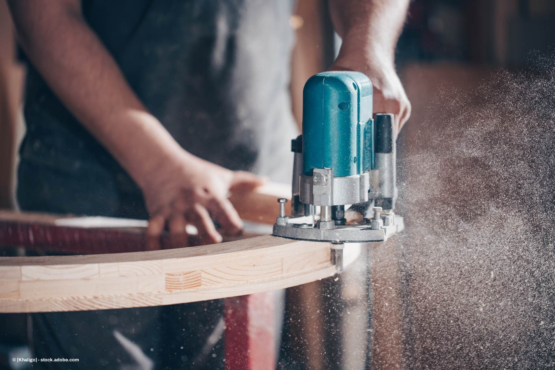 Eine Person bearbeitet mit einer türkisfarbenen Oberfräse ein gebogenes Holzstück und erzeugt dabei Holzstaub.