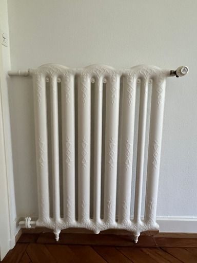 Ein weißer Heizkörper steht auf einem Holzboden neben einer weißen Wand.