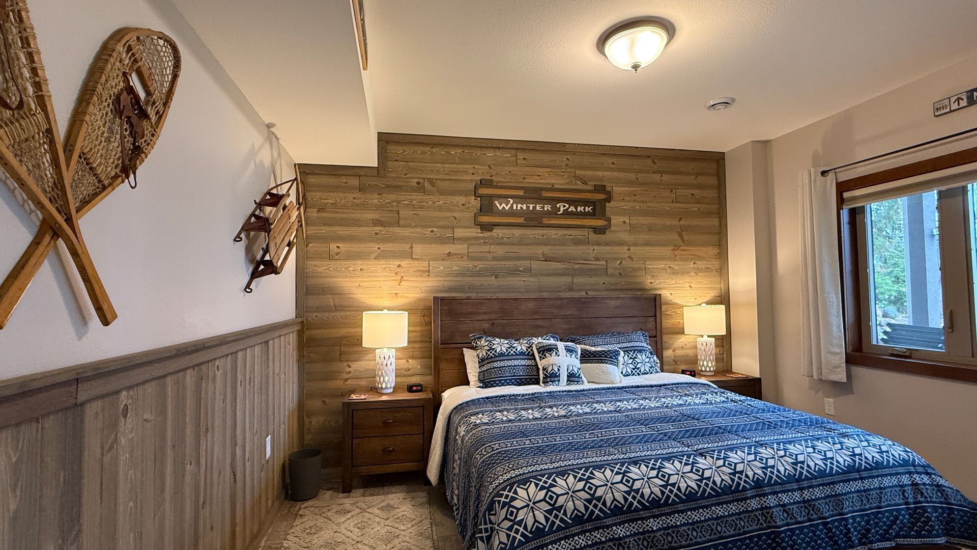 Bedroom with wood paneling, bed, nightstands, and window. Snowshoes hang on the wall.