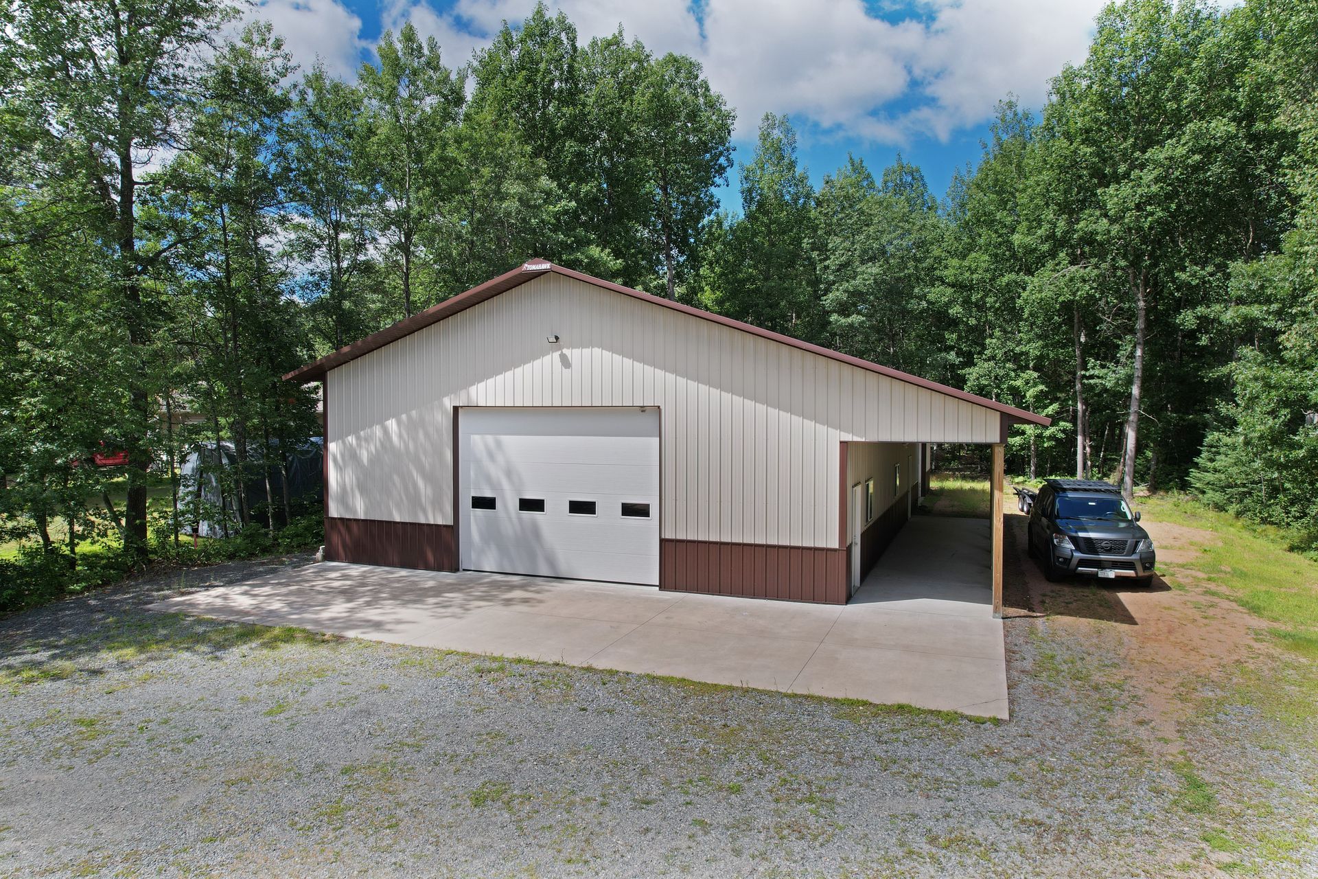 Tan and brown metal building with a garage door and carport; gray gravel driveway; trees in the background.
