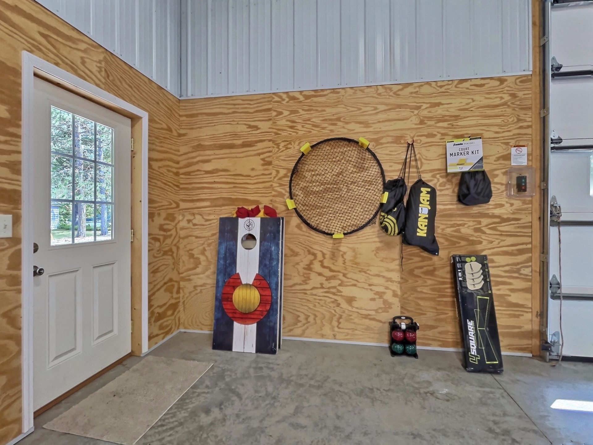 Game room with cornhole, bocce ball, and other outdoor games hanging on a wood-paneled wall, by a white door.