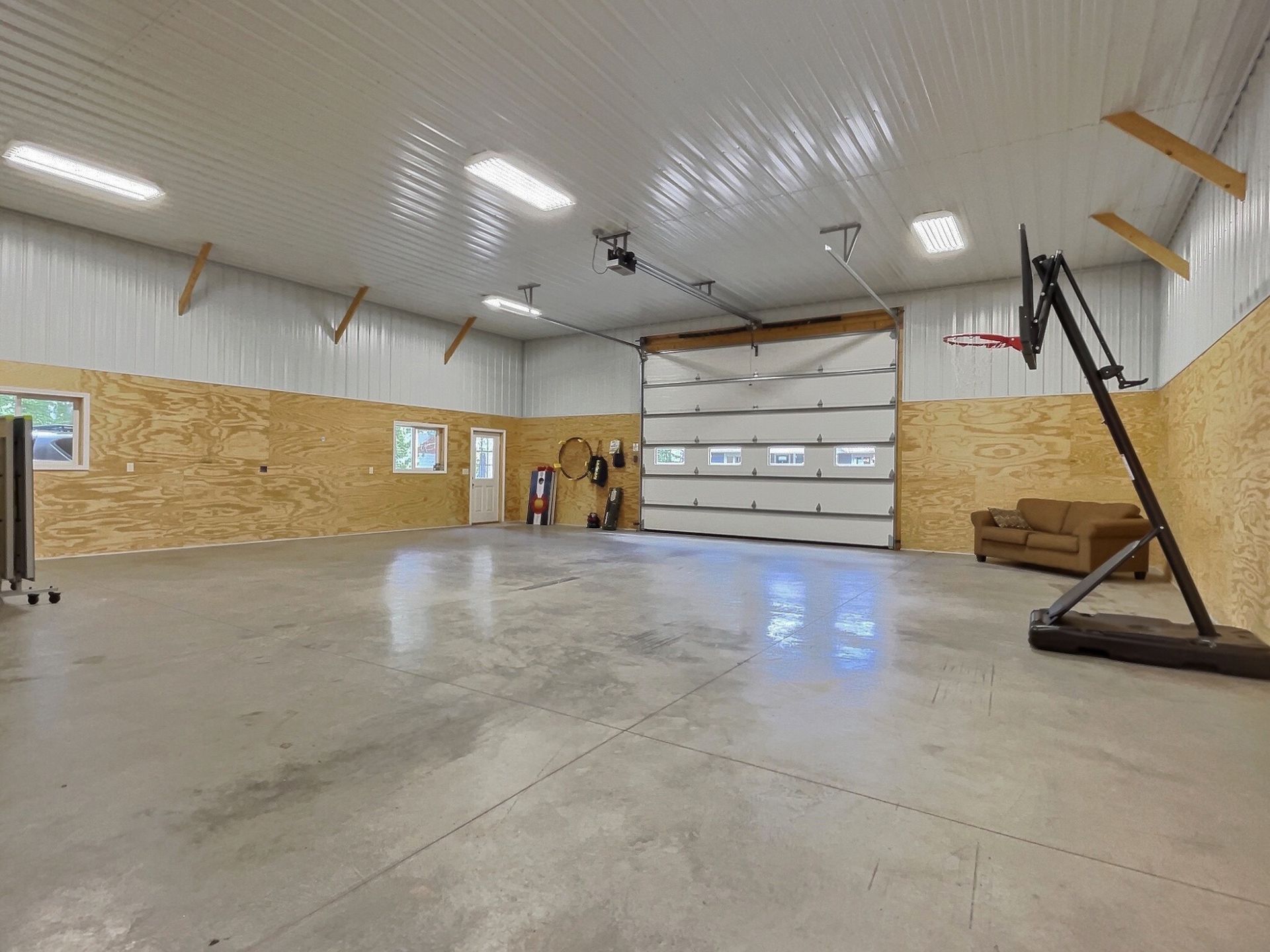 Spacious interior of a garage with concrete floor, basketball hoop, and roll-up door. Walls have plywood and metal sheeting.