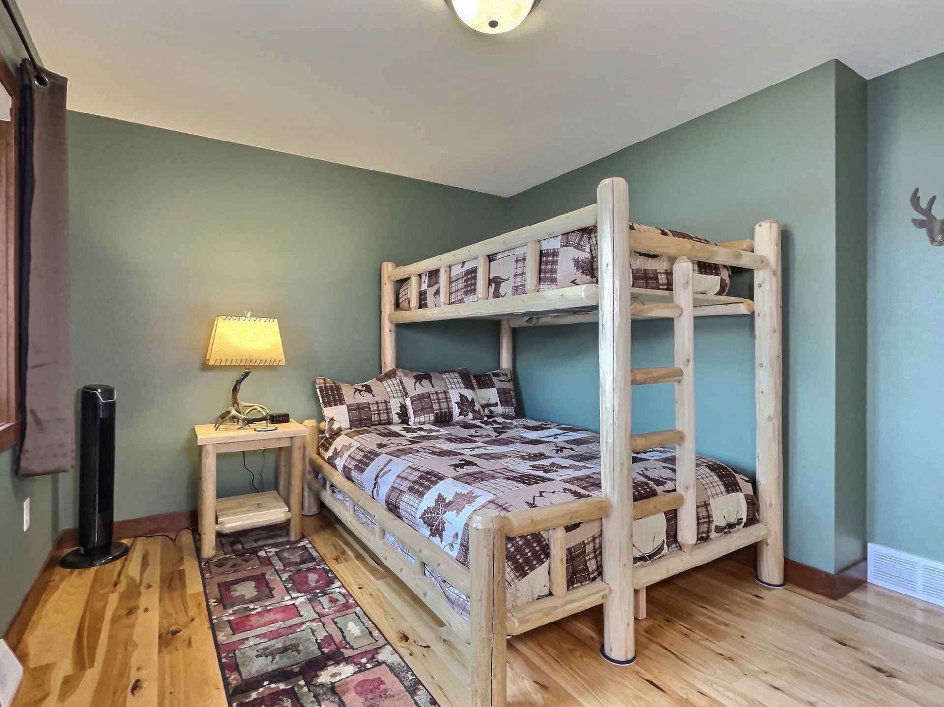 Bunk bed in a bedroom with wooden floors, sage green walls, and a nightstand with a lamp.